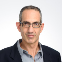 A man with short gray hair and glasses, wearing a dark blazer over a gray collared shirt, poses against a plain light background—ready to tackle the evolving digital fraud battlefield in today's AI-driven cybersecurity landscape.